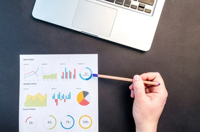 Hand holding pencil reviewing colorful data charts on desk with laptop. Branding agencies attribution methods: four ways